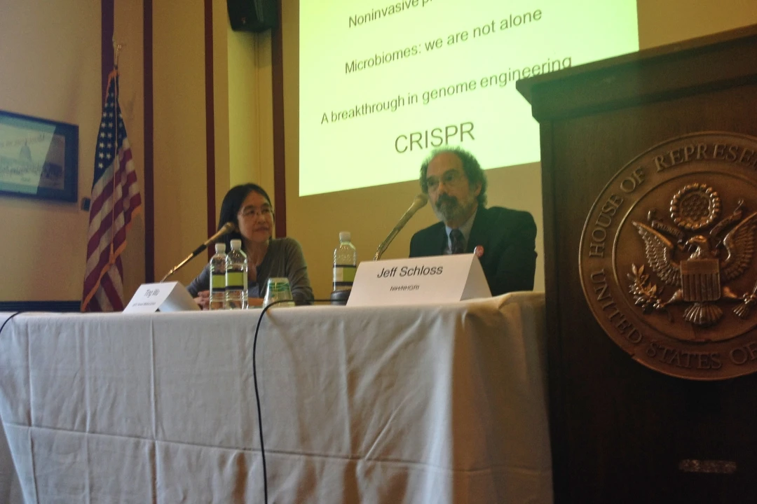 Two people sitting at a table in front of a projection screen. Next to them is a lecture stand with the seal of the United States House of Representatives.