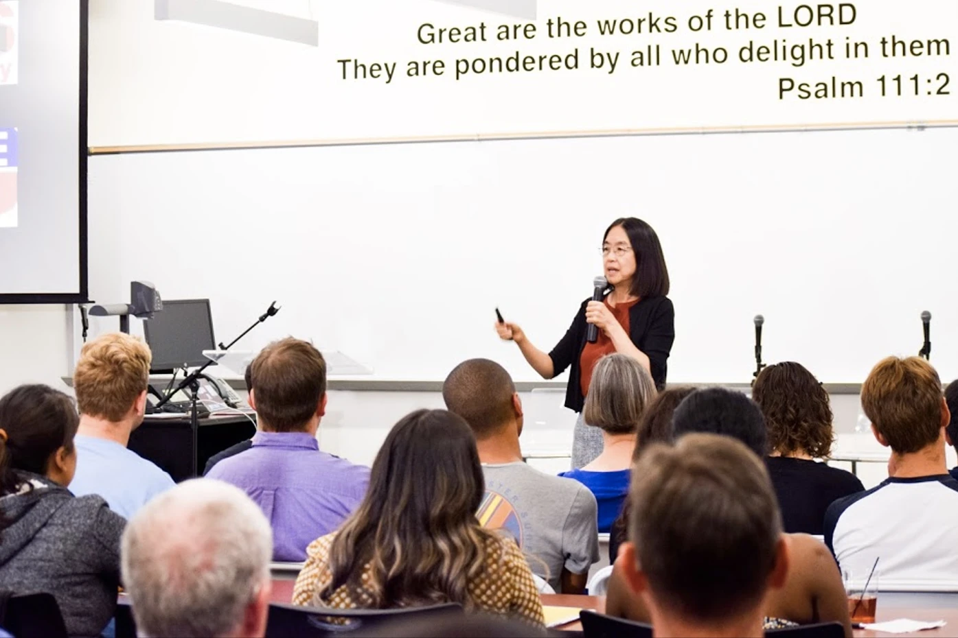 PGED director Ting Wu is holding a microphone and talking with a seated audience. The wall behind her reads “Great are the works of the LORD. They are pondered by all who delight in them. Psalm 111:2”.