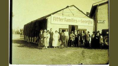 Several families stand in front of a building with a sign saying "Fitter Families for Georgia". Taken at 1924 State Fair in Savannah.