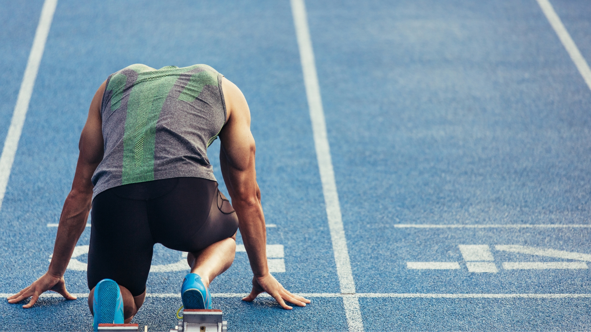 Rear view of a sprinter on a blue running track with his feet in starting blocks.