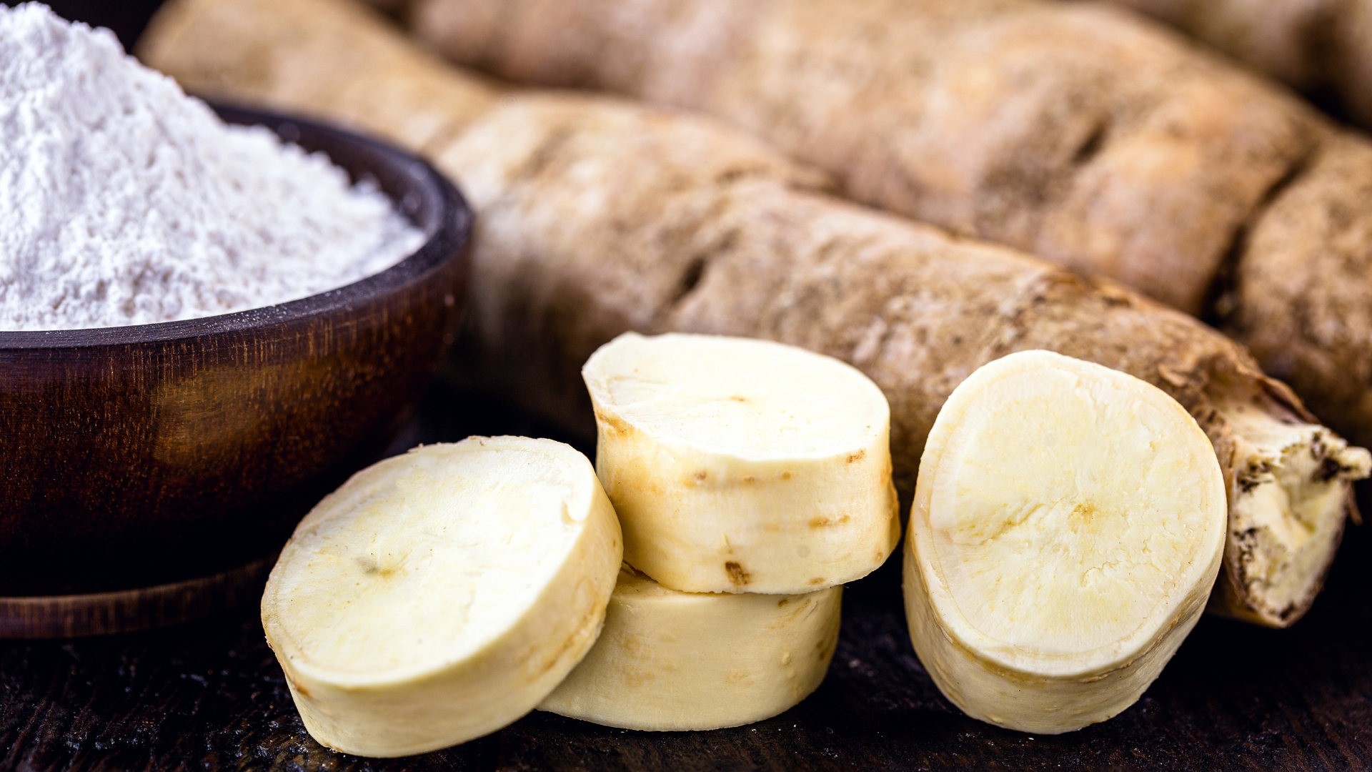 A bowl of white cassava powder next to the raw root and some cut up pieces.