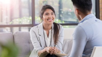 A person smiling and facing another person sitting on a couch talking.